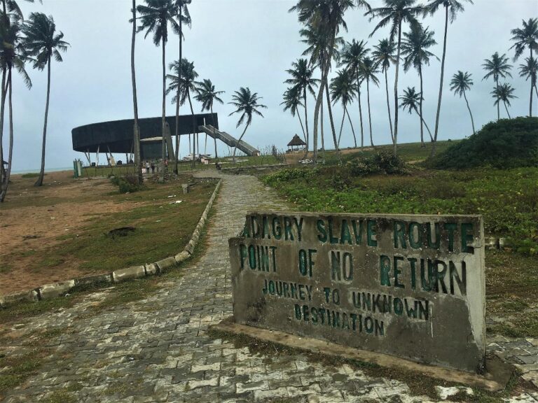 A stone sign reading "Badagry Slave Route, Point of No Return, Journey to Unknown Destination" stands near a path leading towards a beach with palm trees and a dark, modern structure in the distance.